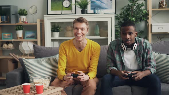 Carefree Male Students Are Playing Video Game on Couch at Home Pressing Buttons on Joystick alt