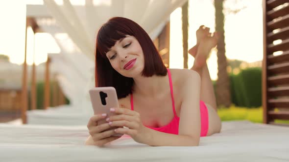 Joyful Young Lady in Pink Swimwear Lying in Gazebo Near the Resort Pool and Using Smartphone alt