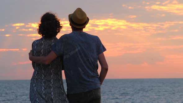 Couple standing on the beach hugging, enjoying sunrise. Silhouette man embraces woman, back view