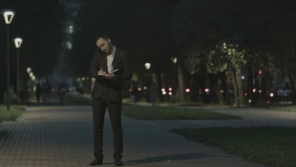 Businessman Using Smart Phone Make a Call While Standing at Park in a City Square, and Make Notes. alt