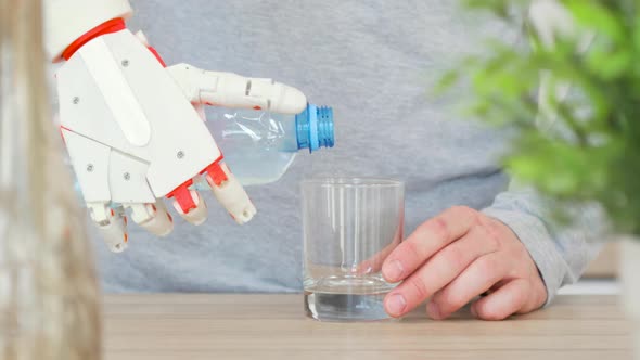 Man with Robotic Prosthetic Hand Is Pouring Water in Glass From the Bottle in the Kitchen alt