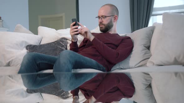 Young attractive man in glasses is sitting on the couch at home and uses his phone. Indoor. alt