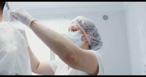 A Nurse Helps a Dentist Put on a White Sterile Suit Tying the Ties of His Back alt
