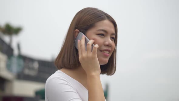 A beautiful Asian woman talks to a cell phone beside the river. alt