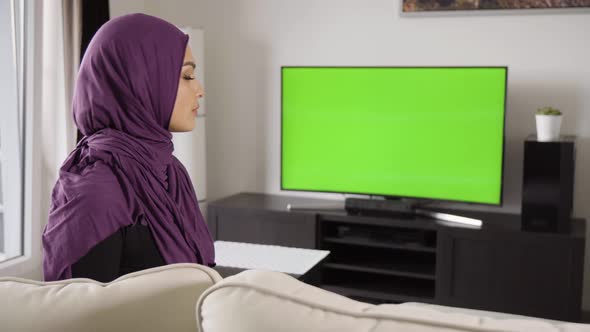 A Young Beautiful Muslim Woman Watches TV with Green Screen and Points at the Camera with a Smile alt