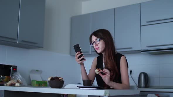 Attractive Young Woman Using Smartphone and Eating at Kitchen