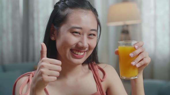Close Up Of Happy Asian Woman Holding A Glass Of Orange Juice And Showing Thumbs Up alt