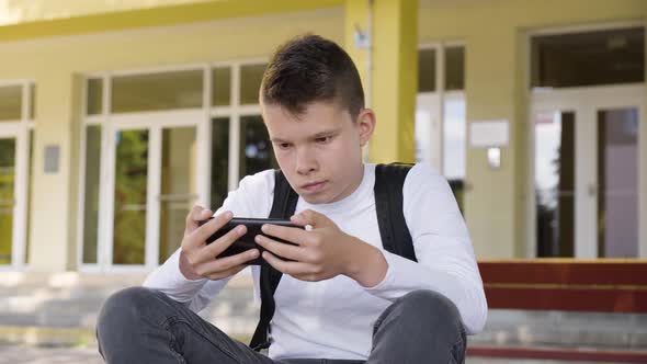 A Caucasian Teenage Boy Looks at a Smartphone As He Sits in Front of School  Closeup