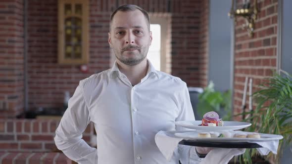 Portrait of Male Waiter Looking at Camera Walking in Slow Motion Holding Tray with Dessert and Salad alt