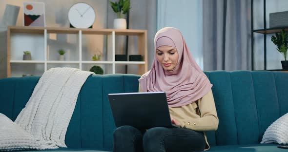 Arabian Woman in Hijab Sitting on Soft Couch in Contemporary Apartment and Uses Computer alt
