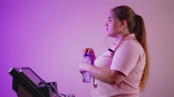 Side View of Perspiring Overweight Young Woman Walking on Treadmill Drinking Refreshing Water alt