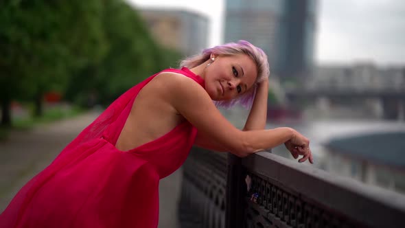 Blonde Woman Is Leaning on Fence of City Embankment and Looking at Camera alt