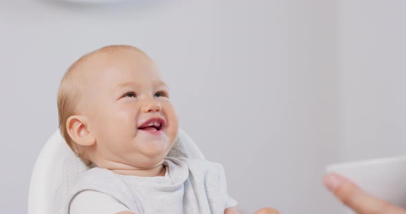Closeup of Young Baby in White High Baby Chair and Father's Hand with a Spoon While He is Feeding alt