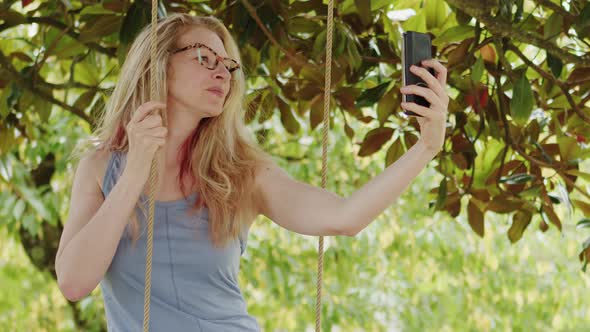 Smiling blonde woman with eyeglasses using smartphone, sitting on the swing