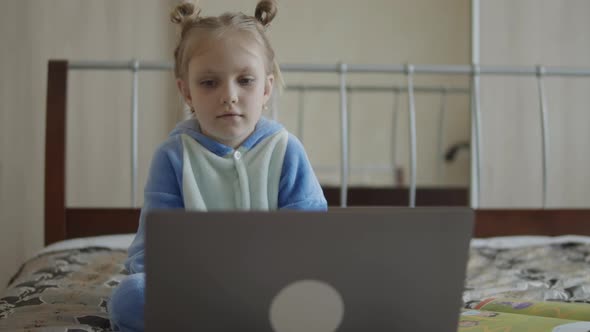 Little Schoolgirl Uses Computer Sitting on the Bed at Home