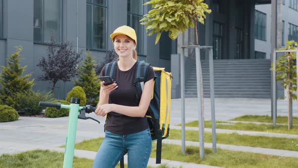 Delivery Woman with Her Electric Scooter Looking at the Phone at the Doors of an alt