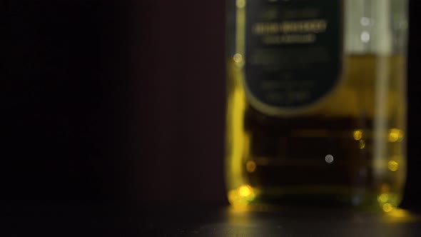 A Man Lifts a Glass of Whiskey From a Table - Closeup - a Bottle in the Blurry Background alt