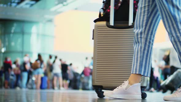 Close-up of Walking Legs in Sneakers, with Suitcase on Wheels, Through Airport Lobby, Against alt