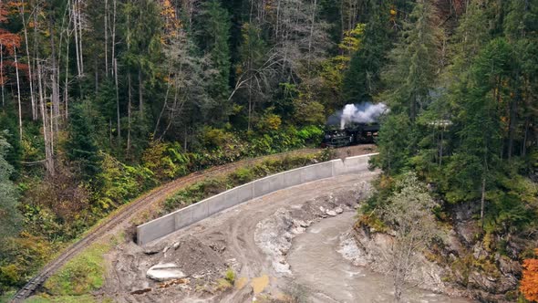 Aerial drone view of the moving steam train Mocanita in a valley along a river, hills covered alt