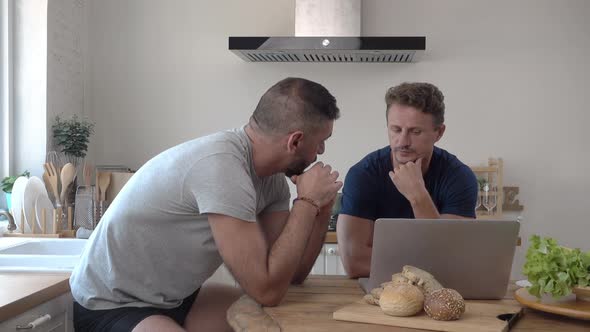 Gay male couple talking seriously while leaning on kitchen table at home