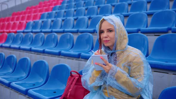 Young Woman in Raincoat with Notepad Pen Sitting on Stadium Bleachers Alone in Rainy Weather alt