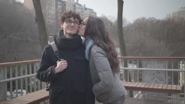 Shy Caucasian Girl in Eyeglasses Kissing Handsome Young Boy on Cheek and Smiling at Camera. Cheerful alt