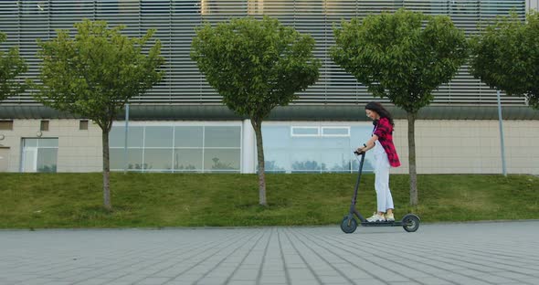 Brunette in Casual Clothes which Riding on Ecological E-scooter Near Big Urban Building alt