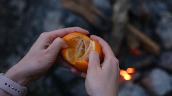Woman hand peeling ripe sweet tangerine, close up above the fireplace in the forest at camping. alt