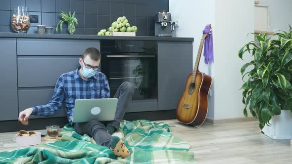 Man in protective medical face mask trying to eat cookies and drink tea during work hours at home alt