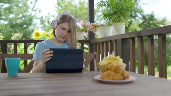 Teenage Girl Sits At Wooden Table In a Summer Cafe In Headphones With Phone alt