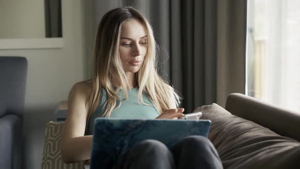 Blonde Woman Sitting on Sofa with Laptop on Knees Using Her Cell Phone alt