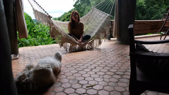 A young woman sitting and relaxing on hammock with a dog sleeping on the floor