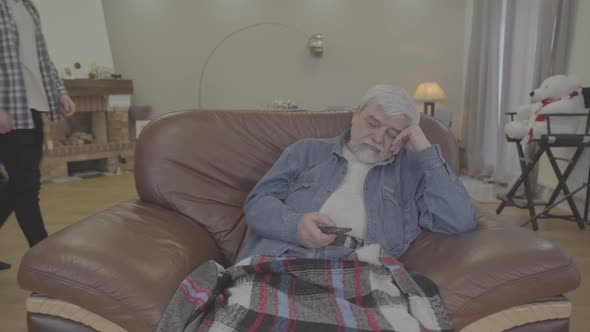 Portrait of Old Caucasian Man Getting Asleep As Watching TV in the Evening at Home. Adult Son Taking alt