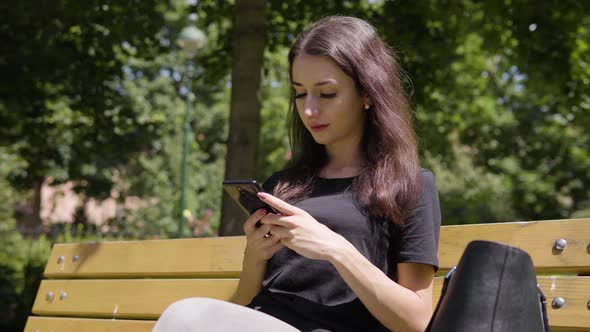 A Young Turkish Woman Works on a Smartphone with a Smile As She Sits on a Bench in a Park alt