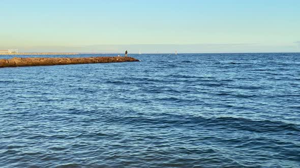 A man is standing on a rock wall fishing in the ocean at sunset. alt