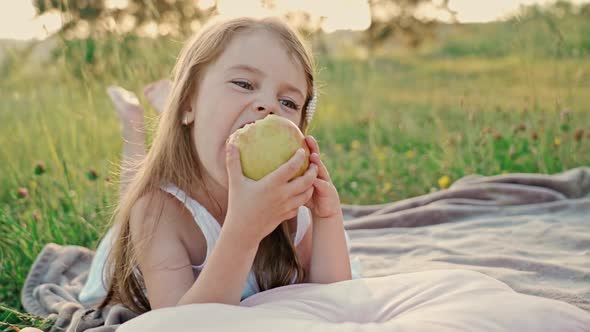Little Cute Girl with Flowing Hair in a White Summer Dress Lying on a Green Lawn Eating an Apple