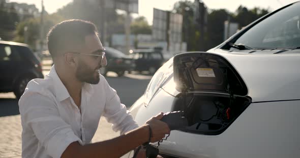 Young Arab Man Recharging White Electric Car at Parking Lot alt