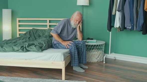 Tired Elderly Man Has Acute Headache While Sitting on Ahe Bed At Home alt