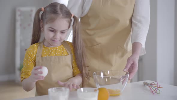 Portrait of Little Concentrated Caucasian Girl Breaking Egg Into Bowl. Cheerful Child in Apron alt