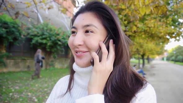 Asian Smling Girl Wearing White Sweater Talking By Smartphone and Walking in Park.