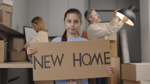 Portrait of Smiling Little Daughter Holding Sign Poster New Home Looking at Camera and Her Parents alt