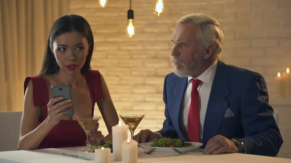 Girl Sitting in Restaurant With Boring Old Man, Chatting on Phone, Bad Date alt