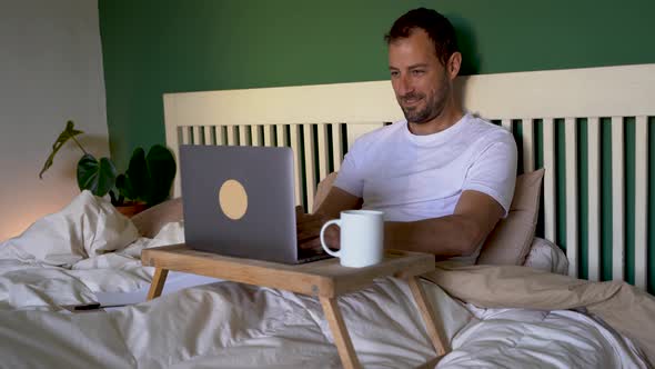 Man sitting in bed working on laptop, drinking coffee