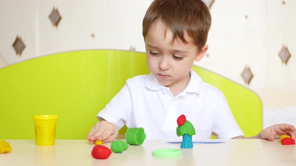 A Child Sitting at the Table, Plays in Colorful Sculptures of Plasticine or Dough for Games alt
