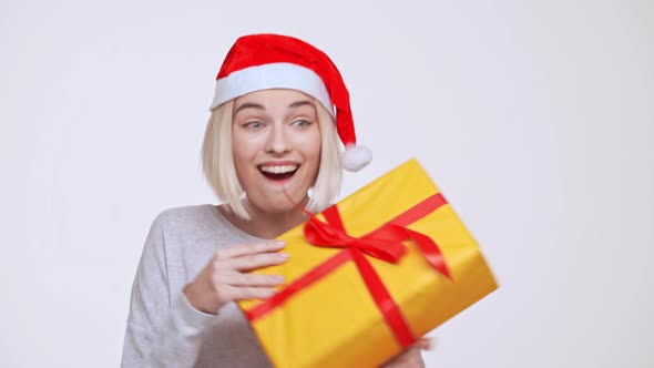 Young Beautiful Blonde Girl in Christmas Hat Suddenly Getting Gift Box White Background alt