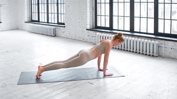 Strong young woman in light beige tracksuit does plank exercise standing on mat alt
