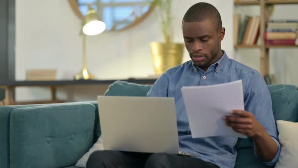 Young African Man with Documents Working on Laptop on Sofa alt