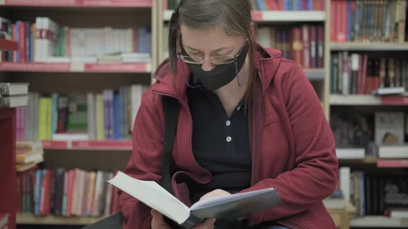 Caucasian Woman Wearing Glasses and Mask Against Virus Reads a Book in Bookshop