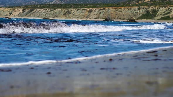 Close-up of Blue Waves Rolling on Sandy Beach with Green Hills at the Background alt