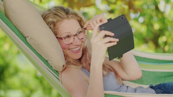 Smiling blonde woman with eyeglasses using smartphone, lying relaxing on the hammock in the garden